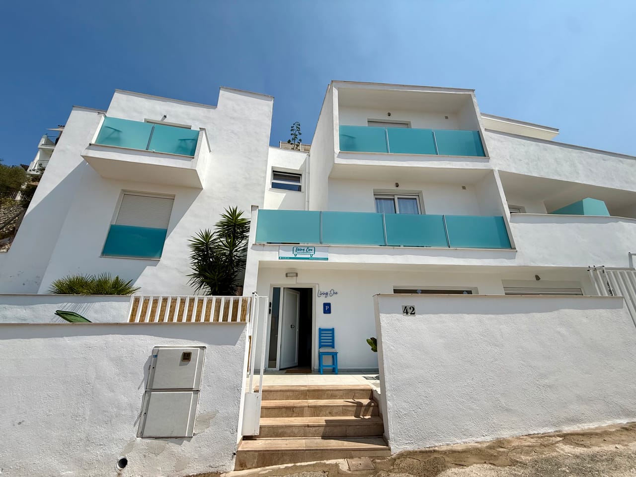 Fachada blanca de Living One con balcones de cristal turquesa bajo el cielo mediterráneo