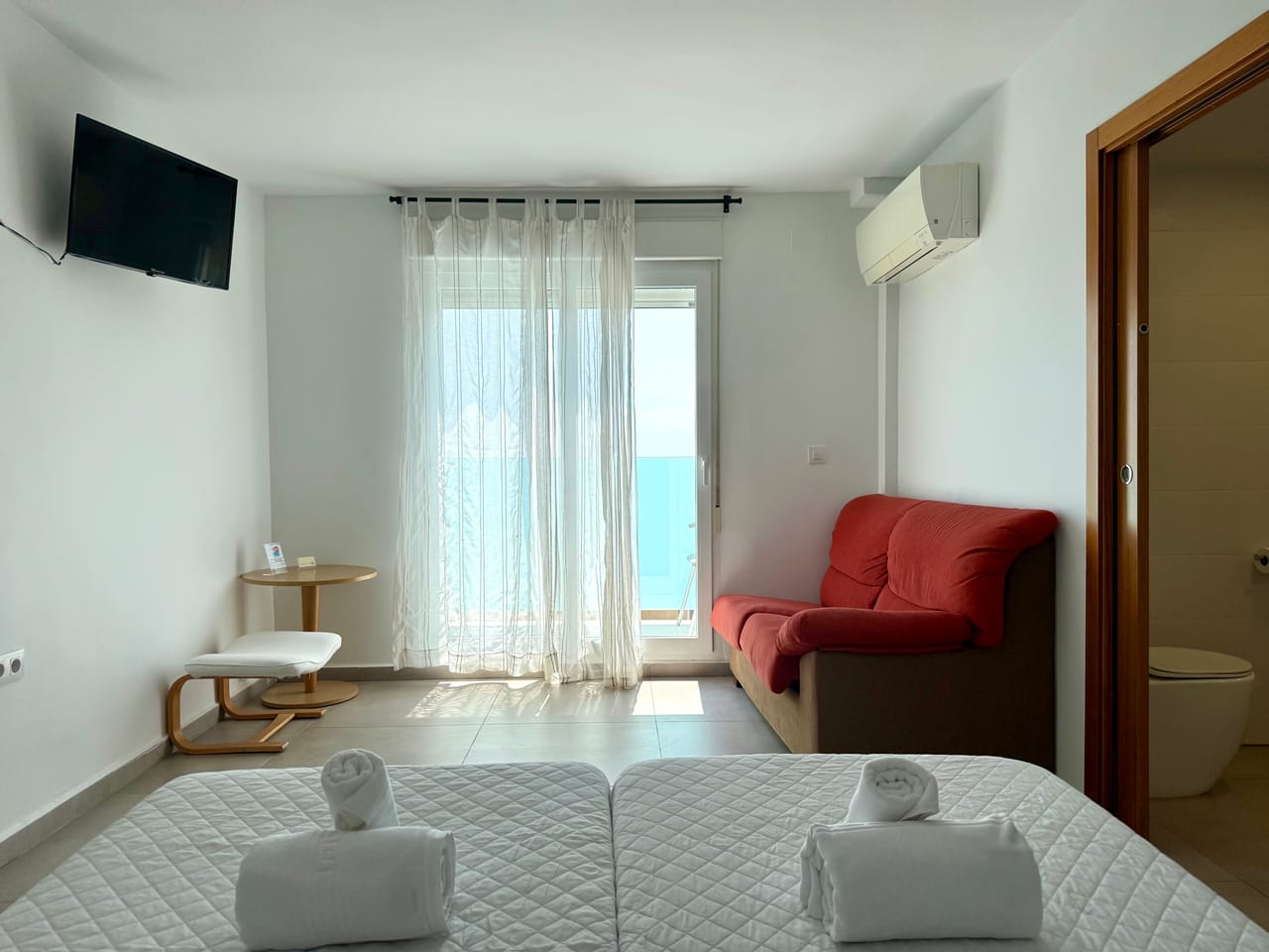 Habitación con sofá burdeos, TV, cortina de lino y baño en suite al fondo