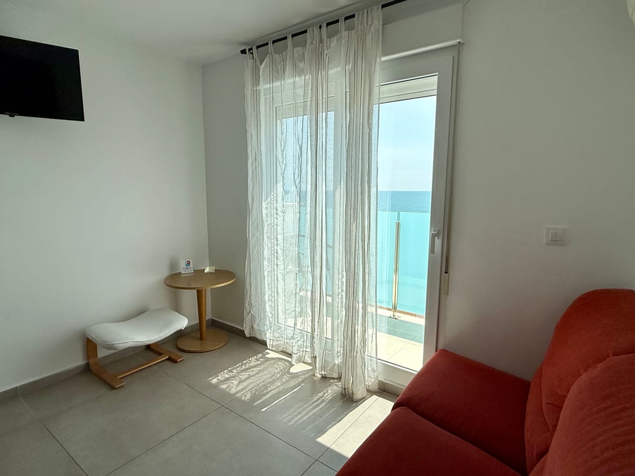 Habitación luminosa con sofá, cortinas de lino y la puerta de la terraza abierta al mar Mediterráneo