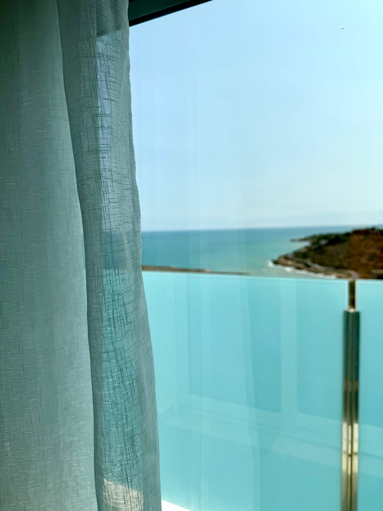Vista al mar Mediterráneo desde la terraza de una habitación, enmarcada por una cortina de lino y la barandilla de cristal turquesa